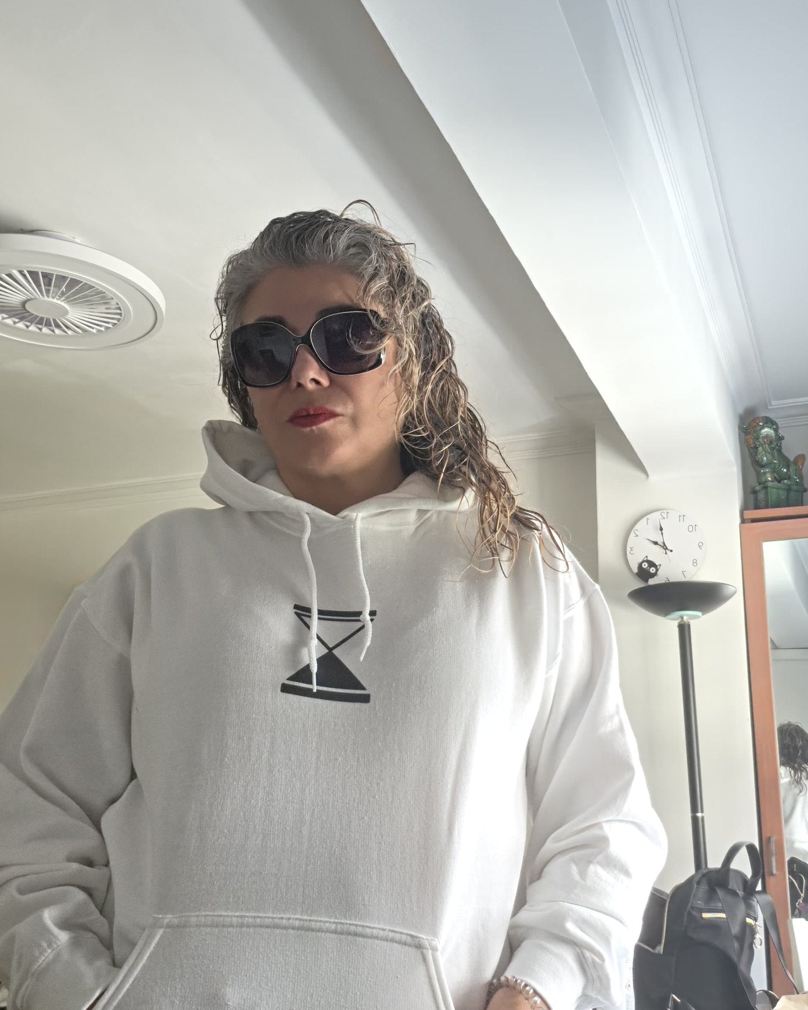 A person wearing a white pullover hoodie with a black geometric hourglass graphic on the chest, styled with dark sunglasses in an indoor setting.